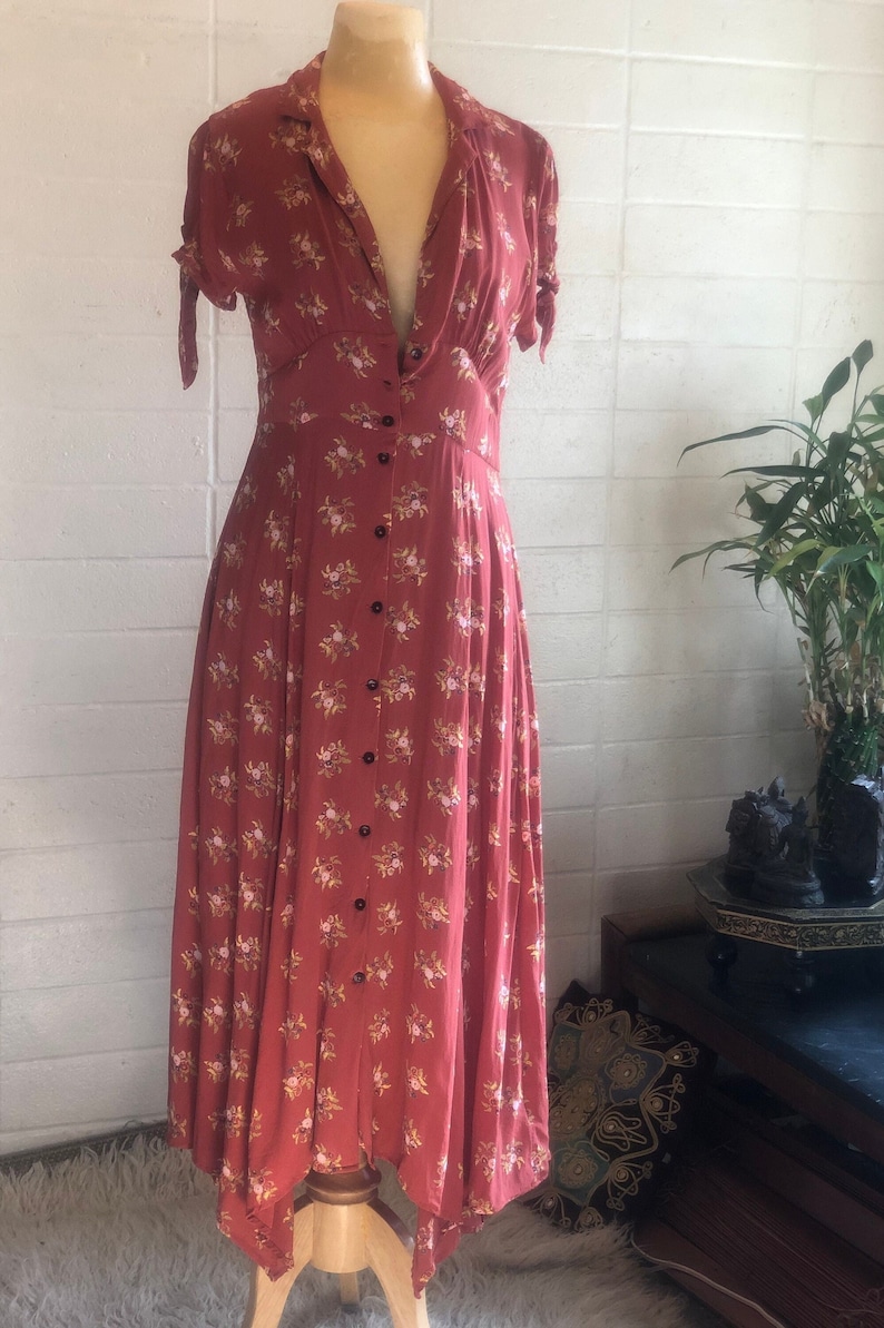 Peut inclure: Une robe maxi de couleur rouille &agrave; imprim&eacute; floral et &agrave; fermeture boutonn&eacute;e sur le devant. La robe a un col en V et des manches courtes avec des liens aux poignets. L'ourlet est asym&eacute;trique, avec un dos plus long et un devant plus court.
