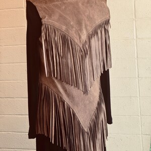 May include: A brown suede vest with long fringe detailing. The vest is layered over a black long-sleeved top. A silver chain necklace is visible at the neckline. The vest has a V-shaped design with fringe cascading down.