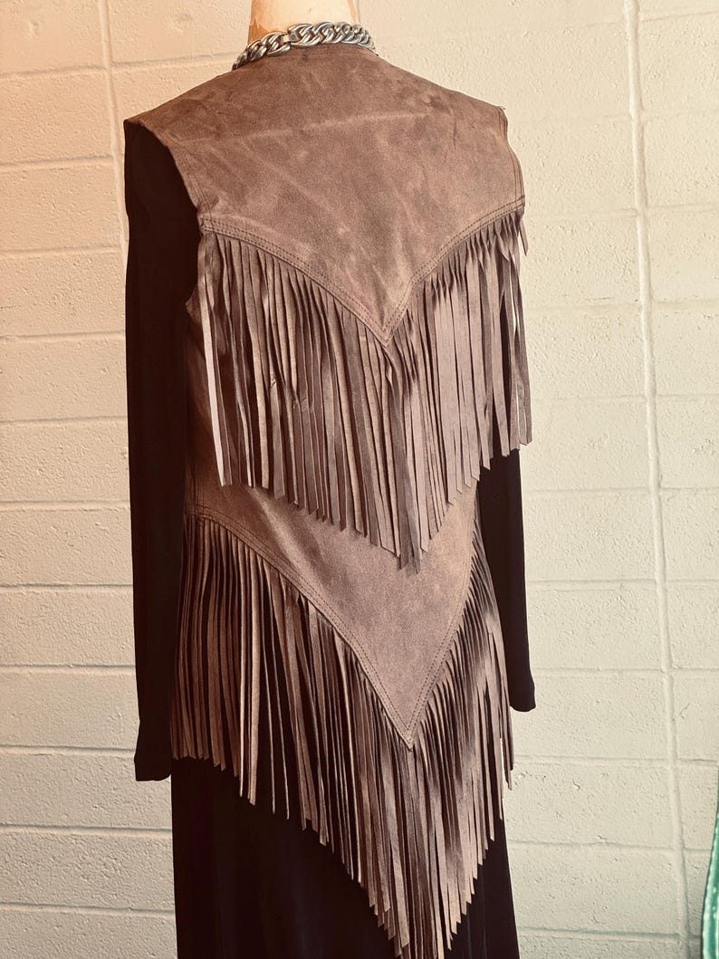 May include: A brown suede vest with long fringe detailing. The vest is layered over a black long-sleeved top. A silver chain necklace is visible at the neckline. The vest has a V-shaped design with fringe cascading down.