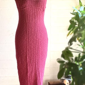 May include: A burgundy ribbed knit dress with a fitted shape. The dress has a scoop neckline, thin straps, and a knee-length hem. A silver chain necklace is worn. The dress is displayed on a mannequin, showcasing its textured fabric.