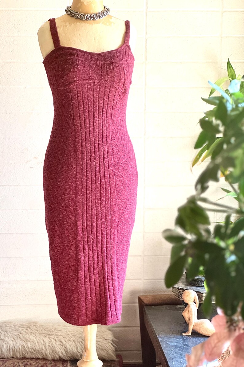 May include: A burgundy ribbed knit dress with a fitted shape. The dress has a scoop neckline, thin straps, and a knee-length hem. A silver chain necklace is worn. The dress is displayed on a mannequin, showcasing its textured fabric.