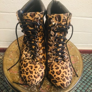 May include: A pair of leopard print boots with black laces and gold-tone eyelets. The boots have a black collar and are displayed on a gold-colored, round table.