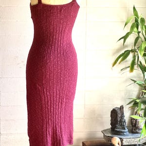 May include: A burgundy-coloured, ribbed knit dress with a fitted silhouette and spaghetti straps. The dress is displayed on a mannequin with a silver chain necklace. The dress is knee-length and has a textured appearance.
