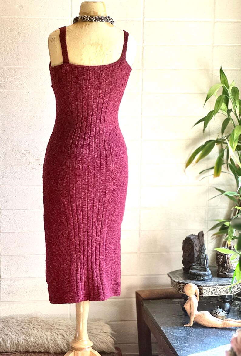 May include: A burgundy-coloured, ribbed knit dress with a fitted silhouette and spaghetti straps. The dress is displayed on a mannequin with a silver chain necklace. The dress is knee-length and has a textured appearance.