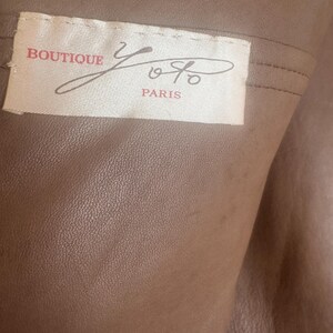 May include: Close-up of a tan leather garment with a label that reads "BOUTIQUE Yoyo PARIS" in red and black script. The leather has a textured surface and is folded, revealing the interior.