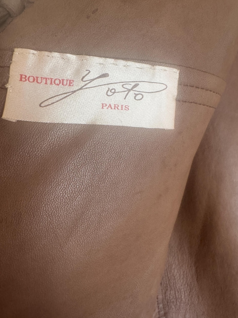May include: Close-up of a tan leather garment with a label that reads "BOUTIQUE Yoyo PARIS" in red and black script. The leather has a textured surface and is folded, revealing the interior.