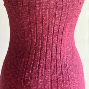 May include: Close-up of a sleeveless, ribbed knit dress in a deep burgundy colour. The fabric has a vertical texture, creating a slimming effect. The dress appears to be form-fitting, highlighting the body's shape. The overall style suggests a casual or semi-formal garment.