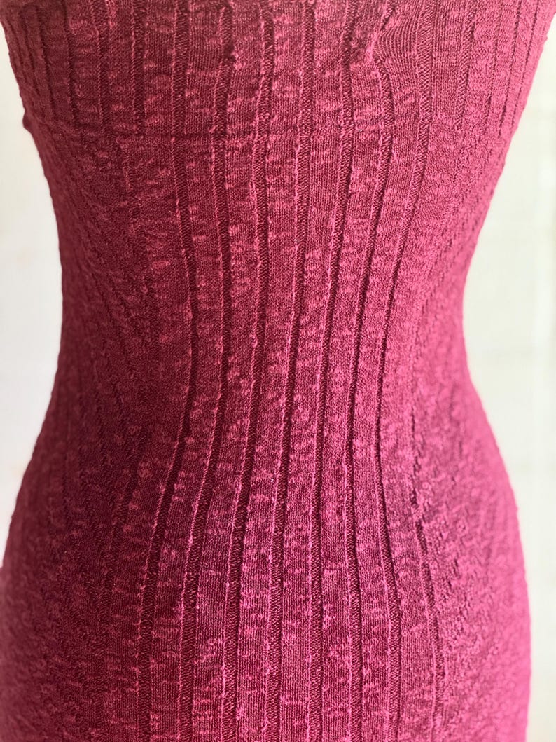 May include: Close-up of a sleeveless, ribbed knit dress in a deep burgundy colour. The fabric has a vertical texture, creating a slimming effect. The dress appears to be form-fitting, highlighting the body's shape. The overall style suggests a casual or semi-formal garment.