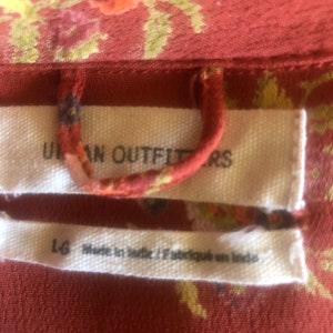 Peut inclure: Une &eacute;tiquette blanche avec le texte "Urban Outfitters" et "L-G Made in India / Fabriqu&eacute; en Inde" sur un tissu floral rouge.