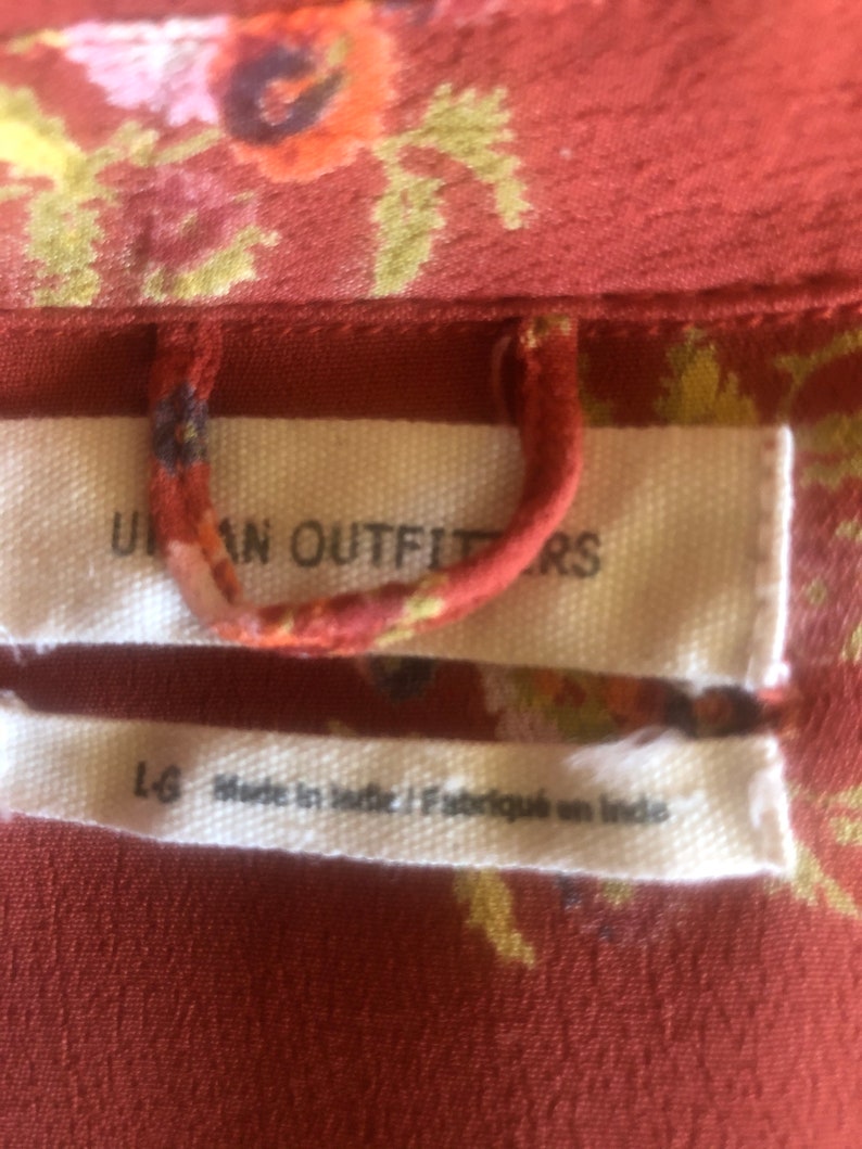 Peut inclure: Une &eacute;tiquette blanche avec le texte "Urban Outfitters" et "L-G Made in India / Fabriqu&eacute; en Inde" sur un tissu floral rouge.