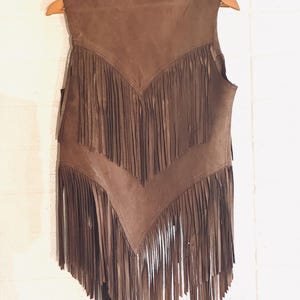 May include: A brown suede vest with long fringe detailing. The vest is sleeveless and hangs on a wooden hanger against a white wall. The fringe is arranged in a V-shape across the chest and along the bottom hem.