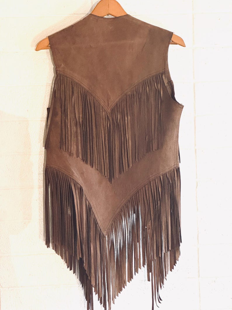 May include: A brown suede vest with long fringe detailing. The vest is sleeveless and hangs on a wooden hanger against a white wall. The fringe is arranged in a V-shape across the chest and along the bottom hem.
