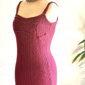May include: A burgundy-coloured, textured knit dress with a fitted silhouette and spaghetti straps. The dress is displayed on a mannequin and paired with a silver chain necklace. The dress has a ribbed texture and a subtle chevron pattern.
