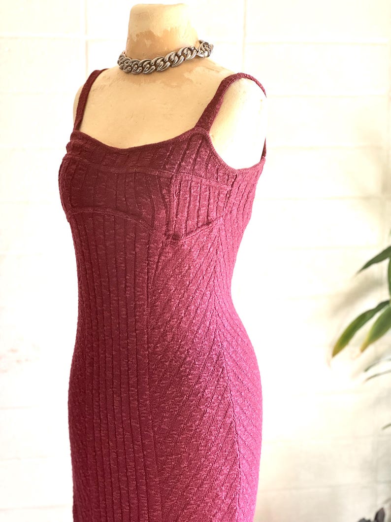 May include: A burgundy-coloured, textured knit dress with a fitted silhouette and spaghetti straps. The dress is displayed on a mannequin and paired with a silver chain necklace. The dress has a ribbed texture and a subtle chevron pattern.