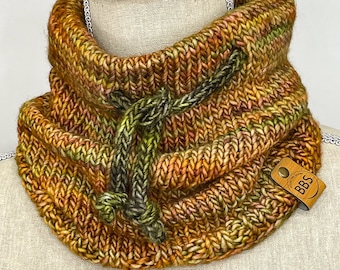 May include: A hand-knitted cowl in shades of brown, orange, and green, with a drawstring closure. The chunky knit neck warmer features a tag that reads "BBS". This cozy accessory is perfect for cold weather.