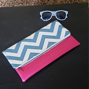 May include: A hot pink clutch with a white and blue chevron pattern flap and a silver zipper. Light blue sunglasses rest above the clutch on a black surface.