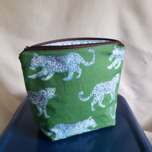 May include: Green fabric cosmetic bag with a repeating pattern of blue leopard print. The bag has a brown zipper and a blue and white patterned interior. The bag is sitting on a blue surface.