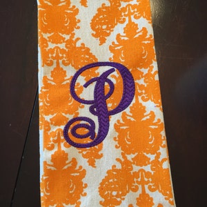 Puede incluir: Una toalla de cocina blanca con un patrón de damasco naranja y una letra "P" bordada en morado en el centro.