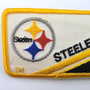 May include: A rectangular embroidered patch featuring a white background with a yellow border. It displays the Steelers logo with yellow, red, and blue stars inside a grey circle, the word Steelers in black text, and a diagonal yellow and black stripe.