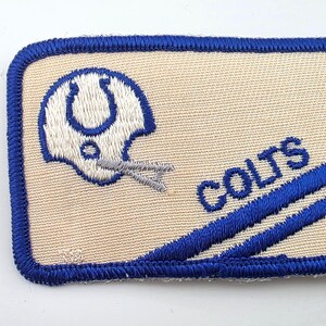 May include: A rectangular embroidered patch with a tan background and a blue border. It features a white football helmet logo with a blue horseshoe, the word COLTS in blue text, and two diagonal blue stripes across the lower right corner.