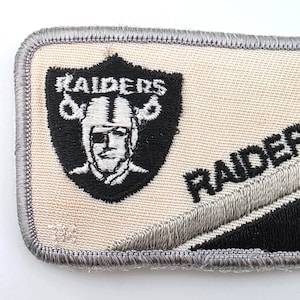 May include: A rectangular embroidered patch featuring a cream-colored background with a grey border. On the left, a black shield displays the Raiders logo with a pirate wearing an eye patch. The word RAIDERS is embroidered in black on the right side.