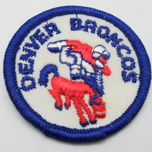 May include: A circular embroidered patch featuring a white background with a blue border. The text Denver Broncos is stitched in blue along the top edge. A red horse with a blue rider is centered on the patch, detailed with white accents.