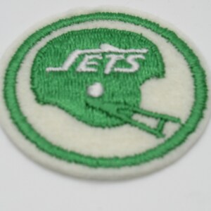 May include: A circular embroidered patch featuring a green football helmet silhouette on a white background. The word JETS is stitched in white letters above the helmet, and the patch is bordered by a thick green stitched circular frame.