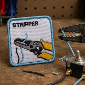May include: A square embroidered patch with a light blue border featuring the word STRIPPER in black text above a graphic of yellow and gray wire strippers cutting a blue wire. The patch is displayed next to actual wire strippers and a toggle switch.
