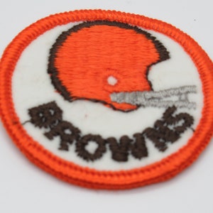May include: A circular embroidered patch featuring a white background with a bright orange football helmet outlined in dark brown. The helmet has a grey facemask. The word BROWNS is stitched in dark brown thread below the helmet. The patch has a thick orange border.