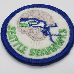 May include: A circular embroidered patch featuring a white background with a blue border. The design includes a light gray football helmet logo with blue and green accents, and the text SEATTLE SEAHAWKS embroidered in green lettering.