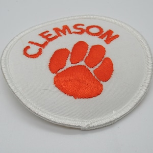 May include: A circular embroidered patch featuring a white background with a bright orange tiger paw print in the center. The word CLEMSON is embroidered in orange capital letters in an arc above the paw print. The patch has a white stitched border.