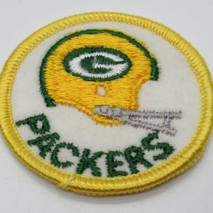 May include: A circular embroidered patch featuring a yellow football helmet with a green G logo, a grey facemask, and the word PACKERS stitched in green thread below the helmet, all set against a white background with a yellow stitched border.