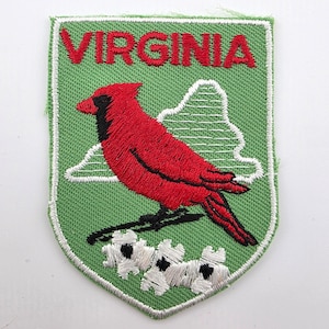 May include: A shield-shaped embroidered patch with a light green background. It features a red cardinal bird with black facial markings, white dogwood flowers at the bottom, a white mountain outline, and the word VIRGINIA in red text.