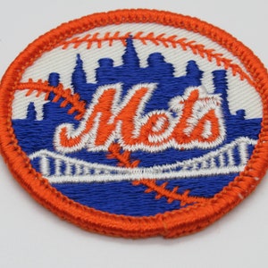 May include: A circular embroidered patch featuring a blue skyline silhouette and a white bridge graphic over a blue background. The word Mets is stitched in orange with a white outline, overlaid on a white baseball pattern with orange stitching.