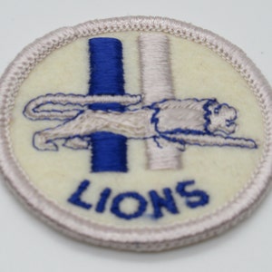 May include: A circular embroidered patch with an off-white background and a light-colored stitched border. It features a blue vertical bar, a white vertical bar, and a lion figure in profile. The word LIONS is stitched in blue at the bottom.