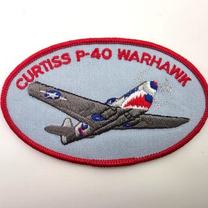 May include: An oval-shaped embroidered patch with a light blue background and a red border. It features a gray Curtiss P-40 Warhawk airplane with a shark mouth nose art design. The text CURTISS P-40 WARHAWK is embroidered in red at the top.