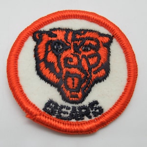 May include: A circular embroidered patch featuring a stylized bear head in orange and dark blue thread on a white background. The bear has an open mouth and is positioned above the word BEARS stitched in dark blue. The edge is finished in orange.