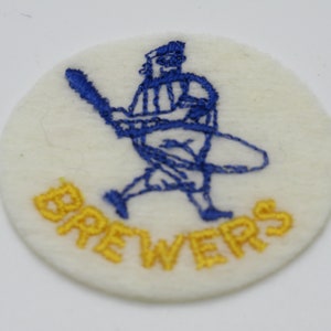 May include: A circular white felt patch featuring an embroidered blue graphic of a baseball player swinging a bat. Below the figure, the word BREWERS is embroidered in yellow thread. The patch has a textured surface and a clean, circular edge.