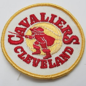 May include: A circular embroidered patch featuring a white background with a yellow border. The text CAVALIERS CLEVELAND is stitched in red around a central yellow circle containing a red silhouette of a basketball player in motion.