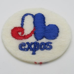 May include: A circular white felt patch featuring an embroidered logo in blue and red. The design includes a stylized blue and red letter M shape above the blue embroidered text that reads expos in a lowercase, rounded, sans-serif typeface.