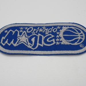 May include: An oval-shaped embroidered patch featuring a blue background with a silver border. The text Orlando MAGIC is stitched in silver, accompanied by a silver basketball graphic and small star accents on the right side of the design.