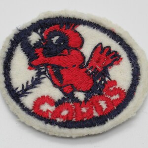 May include: A circular embroidered patch featuring a red bird graphic and the red text "CARDS" on a white background. The design is enclosed within a dark blue stitched border, with a white fabric edge surrounding the entire circular shape.