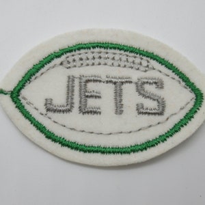 May include: An oval-shaped white felt patch featuring an embroidered football design. The patch has a green stitched border and the word JETS embroidered in silver thread across the center, mimicking the appearance of a football lace pattern.