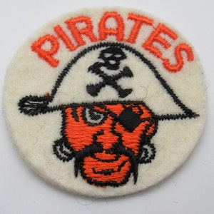 May include: A circular off-white felt patch featuring an embroidered design of a pirate face wearing an eye patch and a hat with a skull and crossbones. The word PIRATES is stitched in orange thread above the hat, with orange and black details.