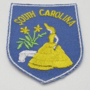May include: A shield-shaped embroidered patch with a light blue background and a white border. The text SOUTH CAROLINA is stitched in yellow at the top. It features a yellow dress, green flowers, and a white architectural element.