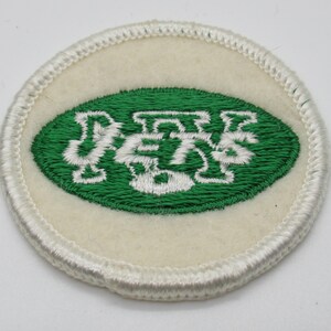 May include: A circular embroidered patch featuring an off-white felt background with a white stitched border. In the center, a green oval contains the white embroidered text JETS, with a football shape integrated into the letter J.