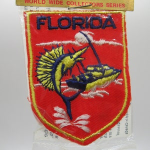 May include: A red embroidered patch featuring the word FLORIDA in dark blue letters at the top. The design depicts a yellow and blue sailfish jumping next to a yellow and blue boat on white waves. The patch is enclosed in a clear plastic bag.