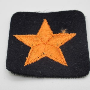 May include: A square black fabric patch featuring a centered, five-pointed orange star embroidered with dense thread. The patch has rounded corners and a visible black stitched border along the bottom edge, showcasing a textured surface.
