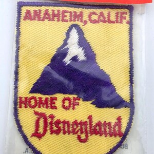May include: A rectangular embroidered patch with a yellow background and a purple mountain peak featuring white snow at the top. The text reads ANAHEIM, CALIF. at the top and HOME OF Disneyland at the bottom in red embroidered lettering.