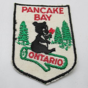 May include: A shield-shaped embroidered patch with a white background and black border. It features the red text PANCAKE BAY above a black bear sitting between two green pine trees. Below the bear is a green banner with the red text ONTARIO.